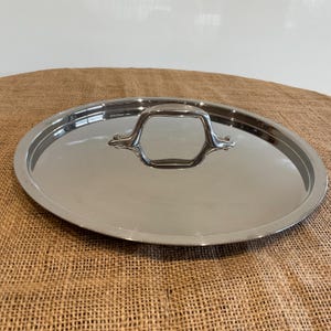 May include: A stainless steel oval pot lid with a handle. The lid is reflective, showing the surrounding environment. The handle is a geometric shape, and is attached to the lid with two small rivets. The lid is sitting on a brown burlap surface.