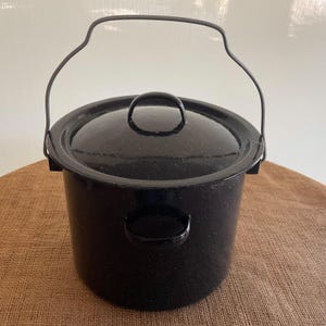 May include: A black enamel stockpot with a speckled finish, featuring a matching lid with a central handle, two side handles, and a tall, arched metal bail handle. The cylindrical pot has a smooth, glossy surface and a wide, flat rim.