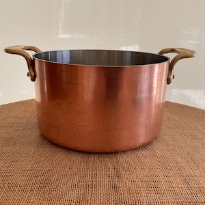 May include: A round copper cooking pot with a polished metallic exterior and a smooth silver-toned interior. The pot features two curved brass-colored handles attached to opposite sides with visible rivets, providing a sturdy grip for lifting.