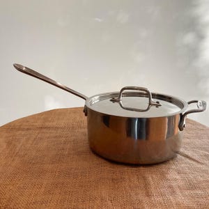 May include: Stainless steel saucepan with a long handle and a lid. The pot has a shiny, reflective surface and a handle on the side. The lid has a loop handle. The pot is sitting on a brown surface.