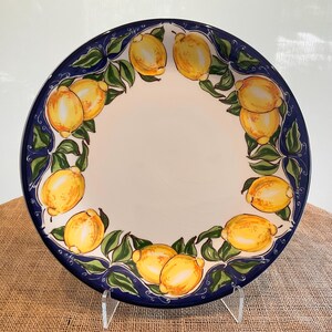 Puede incluir: Un plato redondo de cer&aacute;mica con un centro blanco y un borde azul. El borde est&aacute; decorado con limones amarillos y hojas verdes. El plato se exhibe sobre un soporte transparente.