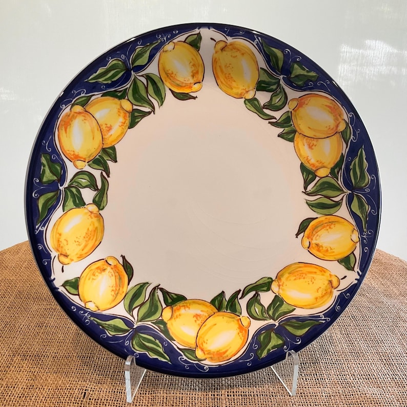 Puede incluir: Un plato redondo de cer&aacute;mica con un centro blanco y un borde azul. El borde est&aacute; decorado con limones amarillos y hojas verdes. El plato se exhibe sobre un soporte transparente.