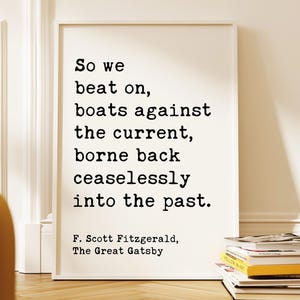 Puede incluir: Un marco blanco con una cita de F. Scott Fitzgerald: "So we beat on, boats against the current, borne back ceaselessly into the past." El texto negro está sobre un fondo blanco. El marco está apoyado contra una pared, con libros en el suelo.
