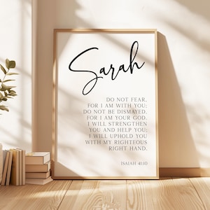 May include: A rectangular wall art print in a light wood frame featuring the name Sarah in large black script at the top, followed by the text of Isaiah 41:10 in a clean black serif font, ending with the citation Isaiah 41:10 at the bottom.