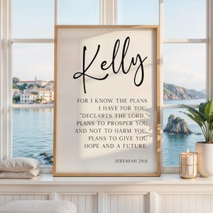 May include: A rectangular wall art print in a light wood frame featuring the name Kelly in large black script at the top, followed by the text of Jeremiah 29:11 in black serif font, centered on a cream-colored background.