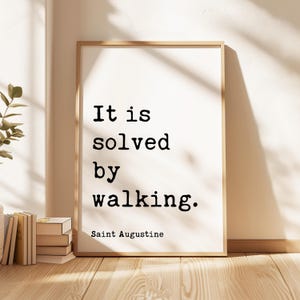 May include: A rectangular wall art print in a light wood frame featuring the black text It is solved by walking. Saint Augustine on a white background. The typography is in a typewriter style font, centered within the frame, with clear lettering.