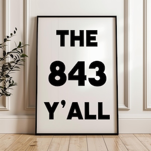 May include: A framed print with the text "THE 843 Y'ALL" in bold black letters on a white background. The frame is black, and the print is displayed against a white wall and wooden floor.