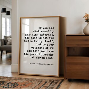 May include: A framed print with a quote by Marcus Aurelius. The text is in black on a white background, set within a light wood frame. The quote reads, "If you are distressed by anything external..." The print is displayed in a well-lit room.