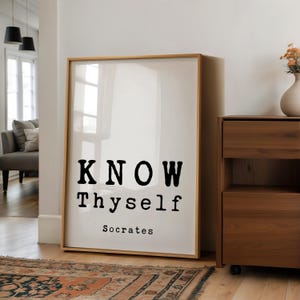 May include: A framed print with the words "KNOW Thyself" in bold black letters, attributed to Socrates. The print has a light wood frame and is displayed against a white wall. The room features a wooden cabinet and a patterned rug.