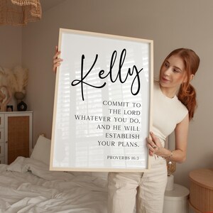 May include: A rectangular wall art print in a light wood frame featuring the name Kelly in large black script at the top, followed by the text: COMMIT TO THE LORD WHATEVER YOU DO, AND HE WILL ESTABLISH YOUR PLANS. PROVERBS 16:3 in black font.