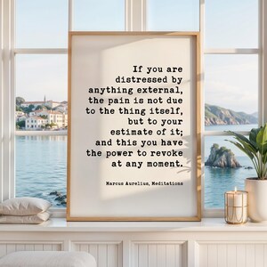 May include: A framed print with a quote by Marcus Aurelius, displayed on a windowsill. The quote is in black text on a white background. The frame is a light brown color. A lit candle and a potted plant are also on the windowsill.