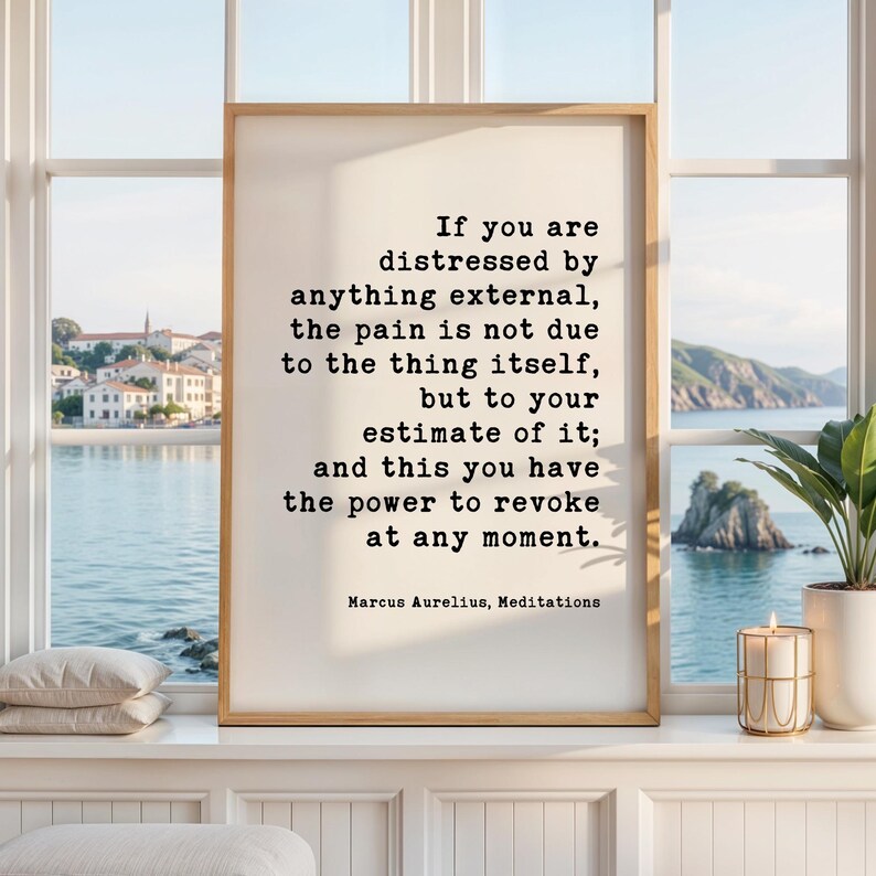 May include: A framed print with a quote by Marcus Aurelius, displayed on a windowsill. The quote is in black text on a white background. The frame is a light brown color. A lit candle and a potted plant are also on the windowsill.