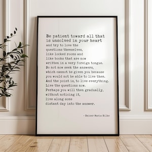 May include: A rectangular black-framed wall art print featuring black typewriter-style text on a white background. The text reads: Be patient toward all that is unsolved in your heart and try to love the questions themselves, signed Rainer Maria Rilke.
