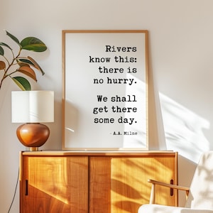 May include: A framed print with the quote "Rivers know this: there is no hurry. We shall get there some day." by A.A. Milne. The print is displayed on a wooden cabinet with a lamp and a plant.