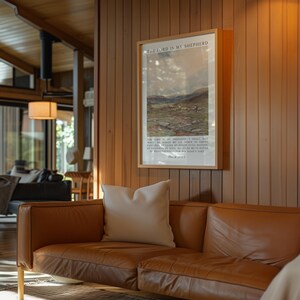 May include: A framed print with the text "THE LORD IS MY SHEPHERD" hangs on a wood-paneled wall. The print features a pastoral scene with sheep. A brown leather sofa with a white pillow is in the foreground.