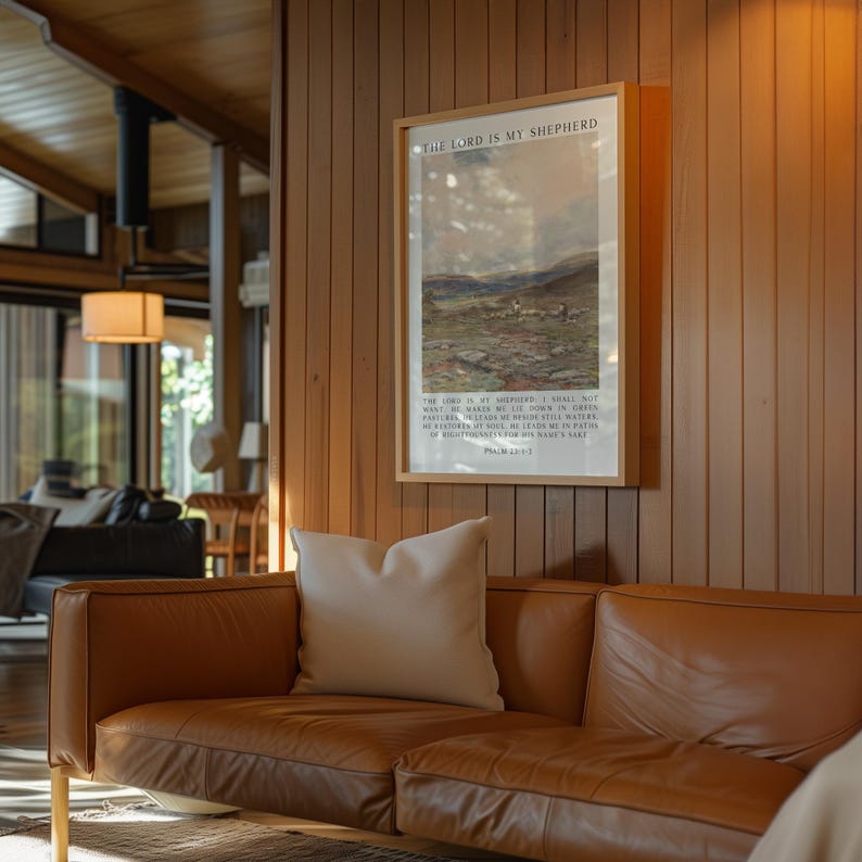 May include: A framed print with the text "THE LORD IS MY SHEPHERD" hangs on a wood-paneled wall. The print features a pastoral scene with sheep. A brown leather sofa with a white pillow is in the foreground.