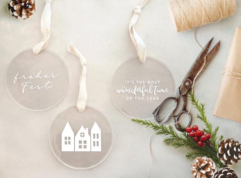 May include: Three round, clear acrylic ornaments with white text and a white silhouette of a house. The ornaments are hanging from white ribbon and are on a light gray surface. The text on the ornaments reads "frohes Fest", "It's the most wonderful time of the year", and a house silhouette.