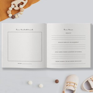 May include: An open baby memory book with text in German, featuring pages for handprints and name details. The book is surrounded by baby shoes, a wooden toy, and other baby accessories.