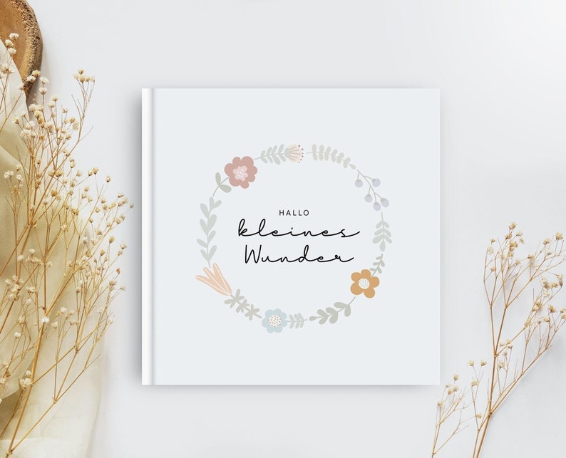 May include: A white book with a floral wreath design and the text "Hallo kleines Wunder" in black.