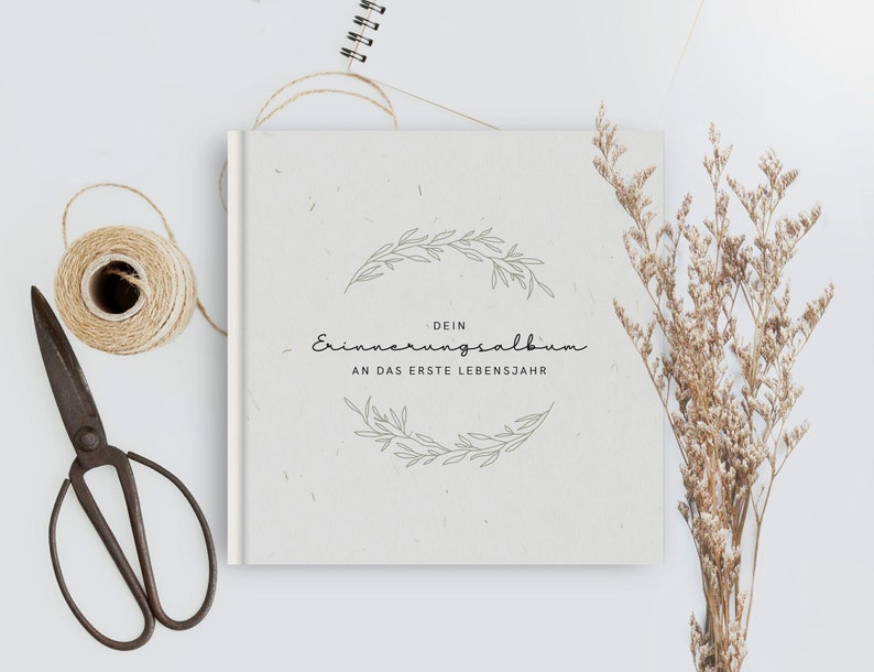 May include: A cream-colored album with the text "DEIN Erinnerungsalbum AN DAS ERSTE LEBENSJAHR" in script font, surrounded by a floral wreath design. A roll of twine, vintage scissors, and dried flowers are arranged around the album.
