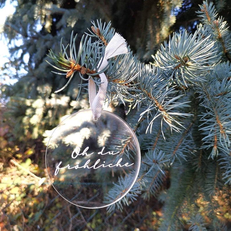 May include: A clear glass ornament with a white ribbon hanging from a pine tree branch. The ornament has the text "Oh du fr&ouml;hliche" written on it.