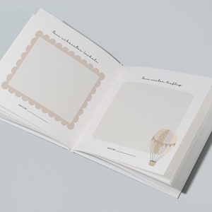 May include: An open baby memory book with two blank pages. The left page has a scalloped frame with the text "Dein wahrer erster Lacher" and the right page has a hot air balloon illustration with the text "Dein erster Ausflug". Both pages have a blank space for a date.