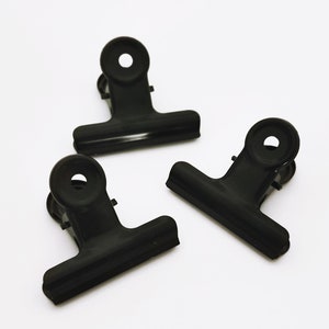 May include: Three black metal binder clips. The clips are closed and have a spring mechanism.