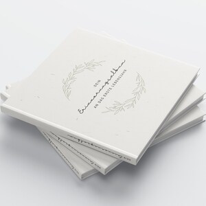 May include: A stack of off-white hardcover books. The top book features the text "DEIN Erinnerungsalbum AN DAS ERSTE LEBENSJAHR" in a handwritten font, encircled by a delicate green leaf design. The books are stacked at a slight angle.