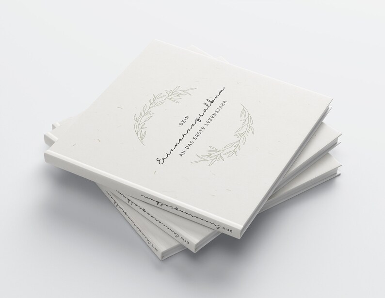 May include: A stack of off-white hardcover books. The top book features the text "DEIN Erinnerungsalbum AN DAS ERSTE LEBENSJAHR" in a handwritten font, encircled by a delicate green leaf design. The books are stacked at a slight angle.