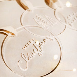 May include: Three clear acrylic Christmas ornaments with white lettering. One ornament says "Merry Christmas", another has a stylized tree design, and the third ornament has a Christmas tree design.
