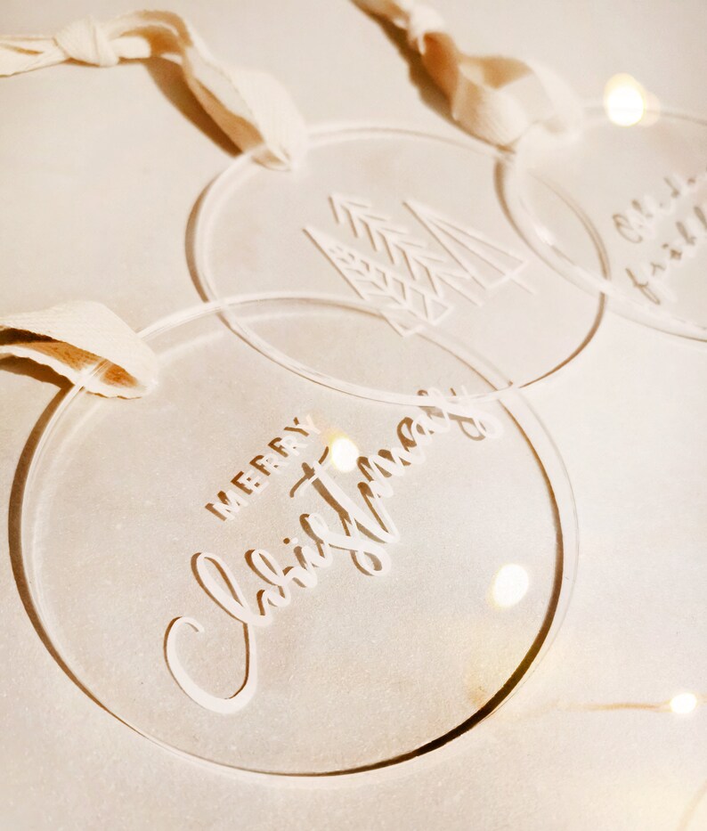 May include: Three clear acrylic Christmas ornaments with white lettering. One ornament says "Merry Christmas", another has a stylized tree design, and the third ornament has a Christmas tree design.