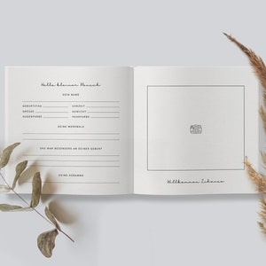 May include: Open baby memory book with the text "Hallo kleiner Mensch" and fill-in-the-blank sections for baby's details. The right page has a photo box and the text "Willkommen Zuhause". Dried plants frame the book.