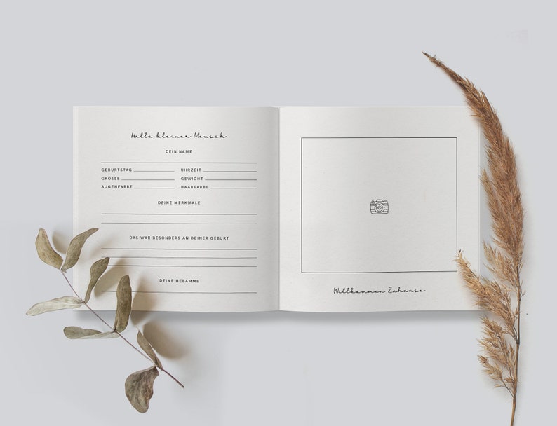May include: Open baby memory book with the text "Hallo kleiner Mensch" and fill-in-the-blank sections for baby's details. The right page has a photo box and the text "Willkommen Zuhause". Dried plants frame the book.