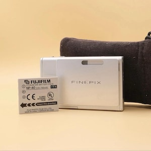 May include: A silver Fujifilm FinePix digital camera positioned next to a rectangular Fujifilm NP-40 lithium-ion battery pack. The battery features black text and a directional arrow, while the camera body displays the FinePix logo.