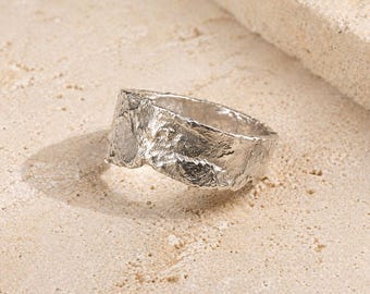 May include: A textured silver ring with an organic, rough-hewn appearance. The ring's surface has a rugged, uneven texture, creating a unique and artistic design. The ring is displayed on a light-colored, textured surface.