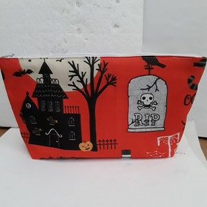 Peut inclure: Pochette rouge à fermeture éclair sur le thème d'Halloween. La pochette présente une silhouette noire d'une maison hantée, un arbre effrayant, une pierre tombale avec un crâne et des os croisés, et les mots "RIP".