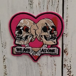 Peut inclure: Patch en forme de cœur rose avec deux crânes brodés se faisant face. Les crânes portent une bannière qui dit "We are as one".