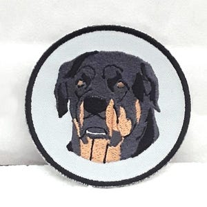 Peut inclure: Un patch brodé représentant la tête d'un Rottweiler en noir et fauve, dans un cercle blanc avec une bordure noire.