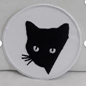 Peut inclure: Patch circulaire blanc avec une silhouette de tête de chat brodée en noir. Le chat a des yeux blancs et un nez blanc.