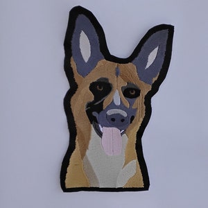 May include: Embroidered patch featuring a German Shepherd dog's face. The patch has a black border, with the dog's fur in shades of tan, gray, and white. The dog's tongue is pink, and its eyes are brown. The patch is on a white background.