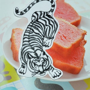 Peut inclure: Broderie de tigre noir et blanc sur fond blanc. Le tigre fait face au spectateur, la gueule ouverte et les dents apparentes. Le tigre est en position accroupie, la queue enroulée autour de son dos.