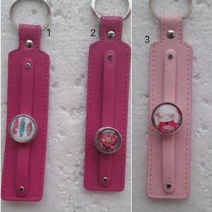 Op de afbeelding: Drie roze en lichtroze leren sleutelhangers met metalen ringen. Elke sleutelhanger heeft een ronde, kleurrijke, decoratieve hanger. De hangers hebben een verenontwerp, een bloemenontwerp en een cartoonfiguur.