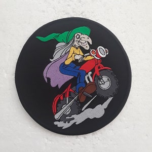 Könnte beinhalten: Ein gestickter schwarzer und grüner Aufnäher mit einer Hexe, die ein rotes Motorrad fährt. Die Hexe trägt einen grünen Hut und ein gelbes Hemd. Das Motorrad hat einen schwarzen Reifen und einen roten Sitz.