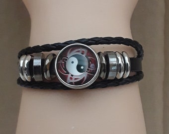 Può includere: Braccialetto in pelle nera con chiusura in metallo argentato e un ciondolo rotondo yin yang con un design rosso e bianco.