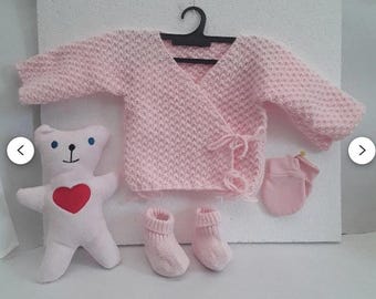 May include: A pink knitted baby cardigan with a wrap-around style and a tie closure. The cardigan is displayed with a pair of pink knitted booties and a pink knitted mitten.