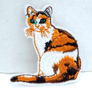 Op de afbeelding: Een calico kat met oranje, zwart en wit bont zit met zijn staart om zijn lichaam gekruld. De kat heeft groene ogen en een witte vlek op zijn borst. De kat is geborduurd op een witte stof.
