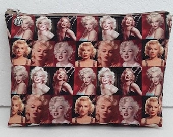 Può includere: Una pochette con cerniera con un motivo a scacchi rosso e nero che presenta un collage di immagini di Marilyn Monroe. La pochette ha una cerniera argentata e un piccolo ciondolo argentato con la parola "LOVE" su di essa.