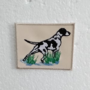 Könnte beinhalten: Gestickter Aufnäher mit einem schwarz-weiß gefleckten Hund in Laufpose, vor beigem Hintergrund. Der Hund ist mit grünem Gras und blauem Wasser dargestellt. Der Aufnäher hat einen beigen Rand.