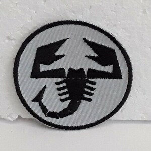 May include: A black and white embroidered patch featuring a scorpion design. The scorpion is in a circle with a white background and a black outline.