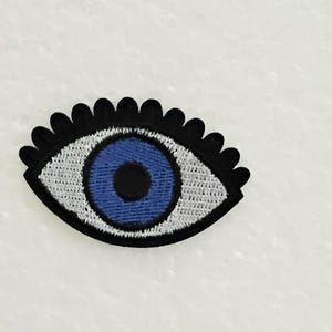 Könnte beinhalten: Ein schwarzer und weißer gestickter Augen-Patch mit einem blauen Auge in der Mitte. Der Augen-Patch ist ein beliebtes Symbol für Schutz und Glück.